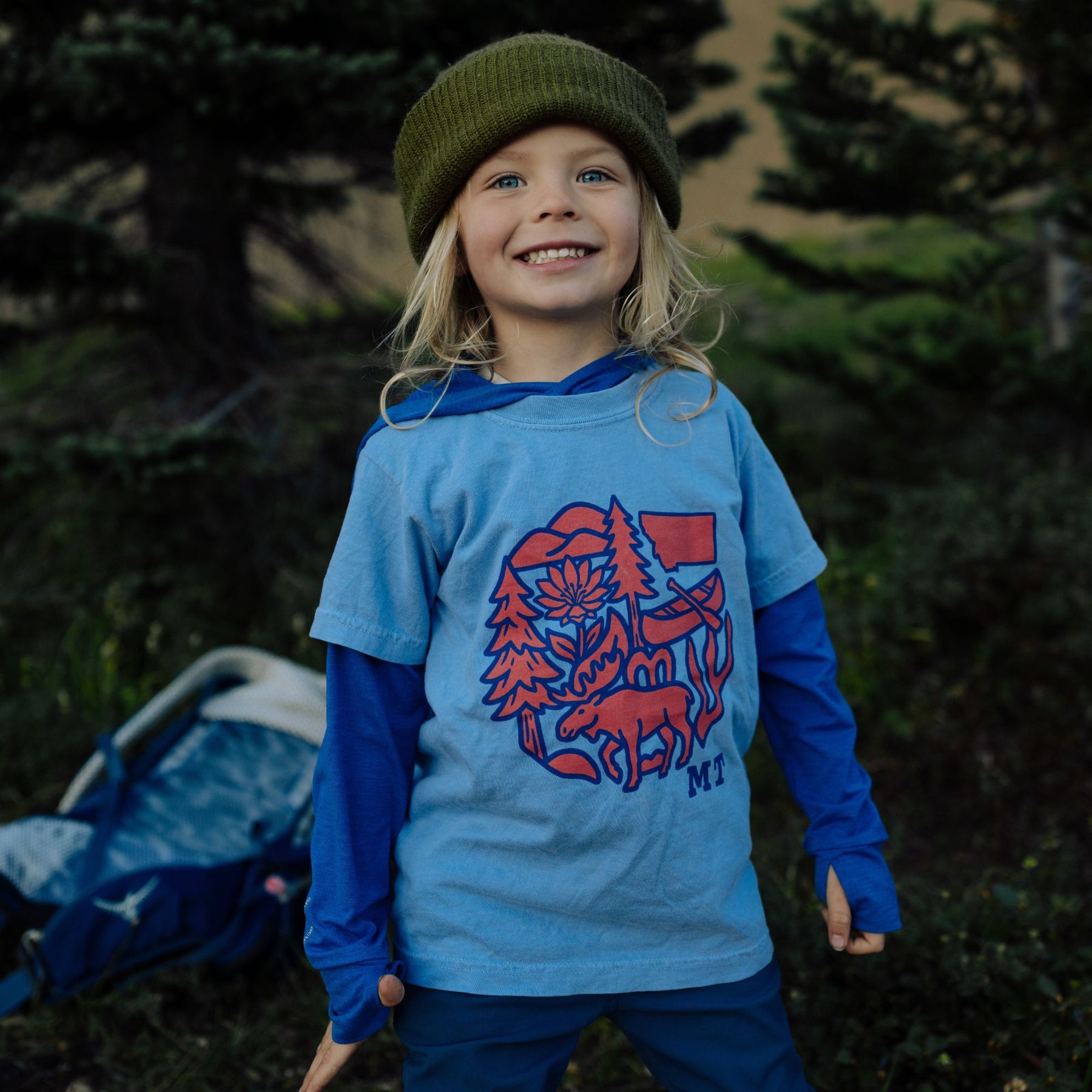 Smiling kid in a green beanie wearing the periwinkle Montana Nature Circle youth tee with a moose and canoe graphic